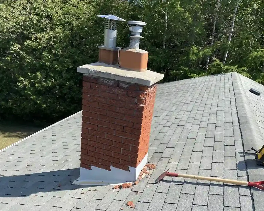 Brick chimney with metal flashing on a grey shingle roof, with tools beside it