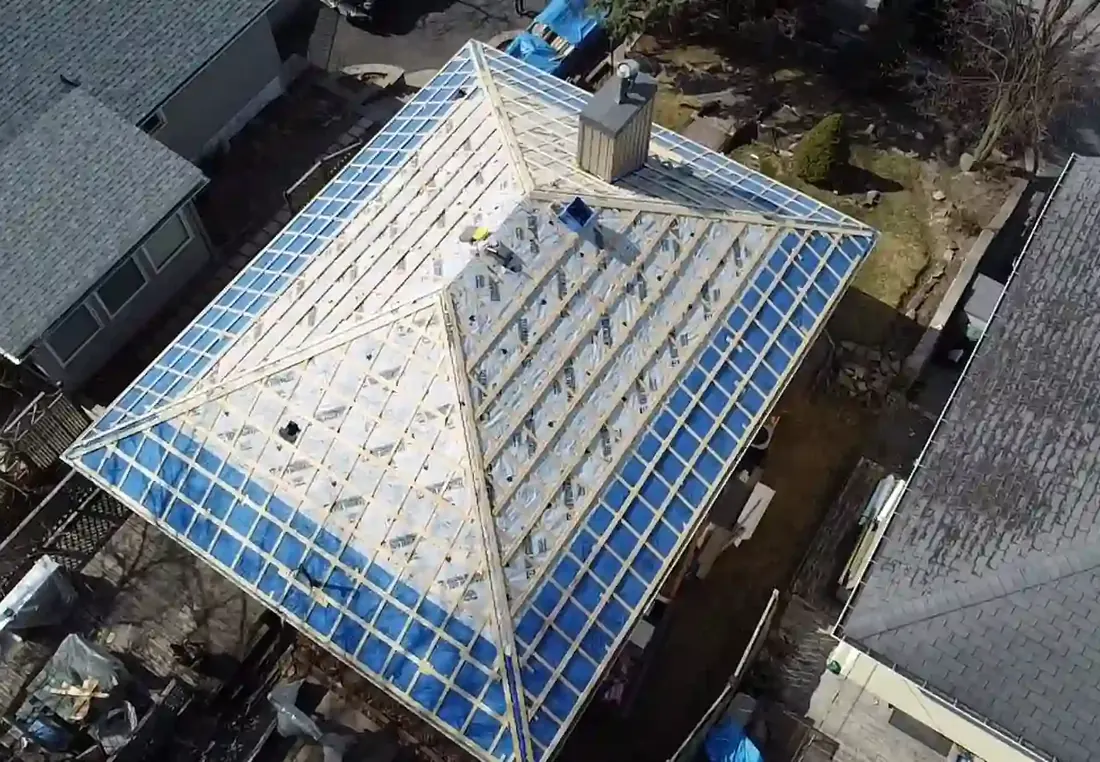 Aerial view of a hip roof stripped to the deck and wrapped in blue and white membrane, with a chimney near the peak